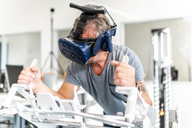 Photo of an employee with VR glasses. He is lying on the Icaros fitness device and holding on to hand attachments.