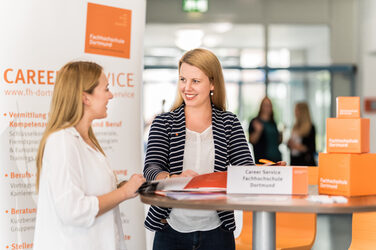 Foto von einer Mitarbeiterin des Career Services der Fachhochschule Dortmund, die an einem Stehtisch im Gespräch mit einer jungen Frau steht. __ <br>Employee of the Career Services of the University of Applied Sciences Dortmund stands at a high table talking to a young woman.