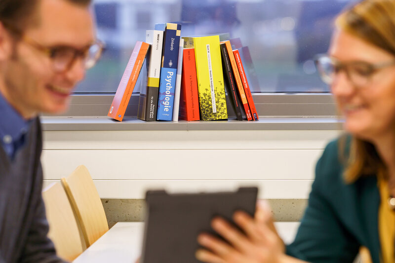 Photo of several books leaning against each other on a windowsill. In the foreground, two indistinctly recognizable people are talking.