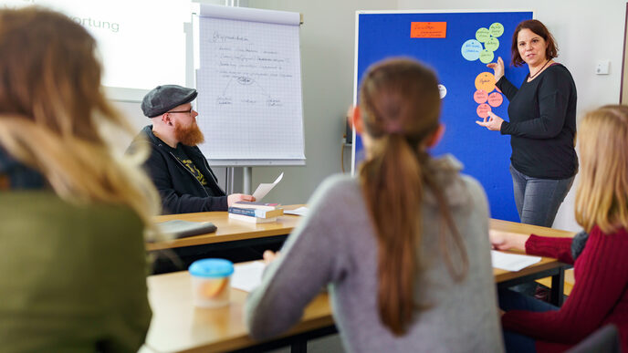 Photo of several participants of the ethnography working group sitting at tables. A woman stands at the front and explains something using a pinboard with several colorful pieces of paper.