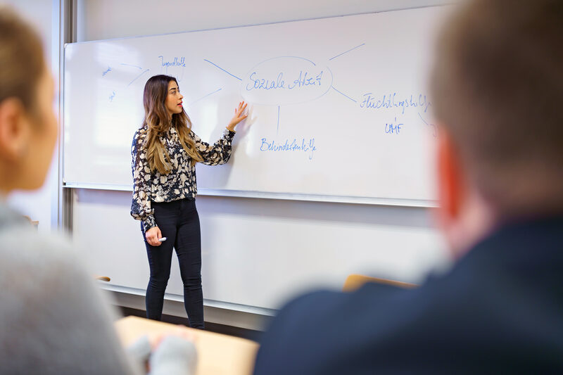 Photo of a young woman standing in front of a whiteboard and explaining a mind map on the subject of social work. Two people listening are out of focus in the foreground.