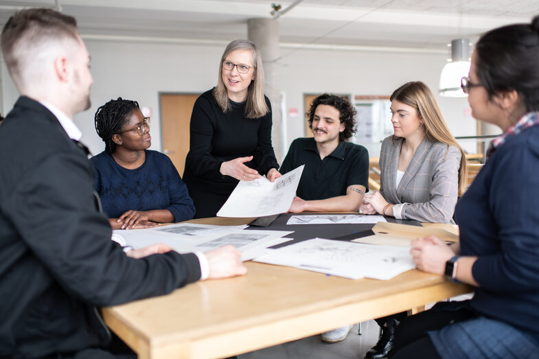 Photo of an employee of the Faculty of Architecture. She is at a table with drawings on it, surrounded by students. She is holding a drawing in her hand and pointing to it with her other hand, smiling as she looks at a student sitting to the left of the picture. __ <br>An employee of the architecture department stands at a table with drawings on it, surrounded by students. She holds a drawing in her hand, which she points to with the other hand, and she smiles and looks at a student who is sitting in the left corner of the picture.