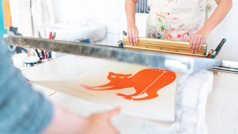 Photo of two people working together to create a screen print of an orange cat. Only the upper body and arms of the people can be seen.