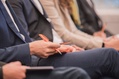 Foto einer Reihe von Menschen, die bei einer Veranstaltung sitzen. Ein Mann hält Kugelschreiber und Material in der Hand. __ Row of people sitting at an event, man holding pen and material in hand.