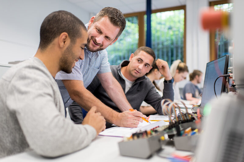 Foto aus dem Labor für Elektronik und Automation. Ein Professor hilft zwei Studierenden an Arbeitsplätzen mit elektronischen Versuchsaufbauten weiter. Im Hintergrund weitere Studierende.