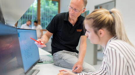 Foto einer Studentin im Labor für Elektronik und Automation, die an einem Computer sitzt. Neben ihr sitzt ein Mitarbeiter auf dem Tisch und zeigt ihr mit einem Stift etwas auf dem Bildschirm.