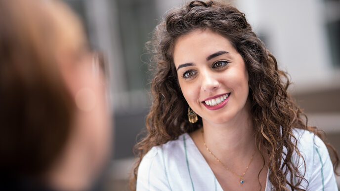 Closeup-Aufnahme einer jungen Frau mit dunklen lockigen Haaren, die eine Frau die ihr gegenüber sitzt anstrahlt.__Closeup of a young woman with dark curly hair smiling at a woman sitting across from her.