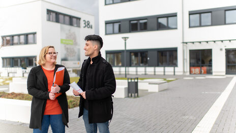 Foto einer Mitarbeiterin, die mit einem Schüler vor einem Campusgebäude steht. Sie hält eine orangene Mappe unterm Arm, er hat einen Flyer in der Hand.