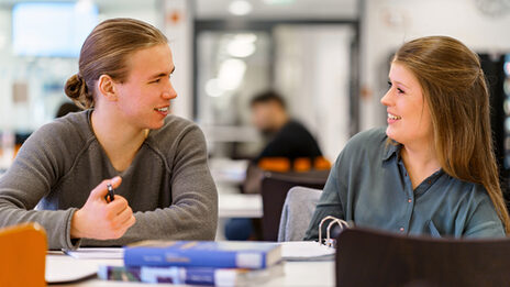 Foto von einer Studentin und einem Studenten nebeneinander an einem Tisch, sie lachen sich an. Auf dem Tisch liegen Bücher.__Student sit next to each other at a table, they laugh at each other. Books are on the table.<br><br>