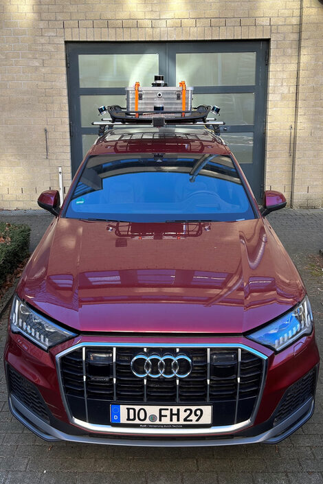 A black Audi Q7 is parked in front of a building. The car has a large roof rack on which a silver box with orange straps and several sensors are attached. The vehicle is shown head-on and is parked on a paved parking lot in front of a building and a gray door with several windows.