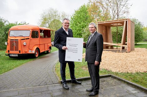 Zwei männlich gelesene Personen stehen vor der Holzkonstruktion eines dreidimensionalen Würfels. Sie halten eine Urkunde in den Händen.