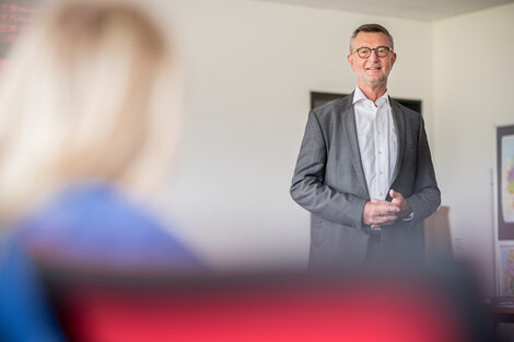 Foto eines Mitarbeiters, der im Raum steht und ein Seminar mit dem Thema Existenzgründung hält. Links im Bild ist sehr unscharf eine sitzende Person zu erkennen.