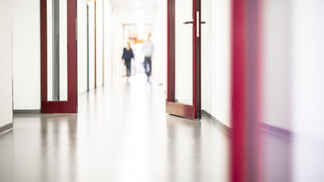 A photo of a long corridor with a red door in the foreground. Two people are walking along the corridor.