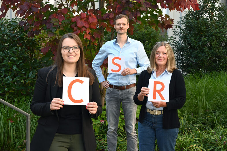 Group picture of CSR Office teams: three people each hold up a sheet of paper with an orange letter on it. From left to right, this is how CSR is read.