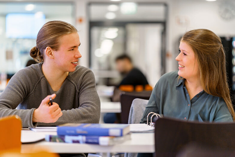 Foto von einer Studentin und einem Studenten nebeneinander an einem Tisch, sie lachen sich an. Auf dem Tisch liegen Bücher.__Student sit next to each other at a table, they laugh at each other. Books are on the table.<br><br>