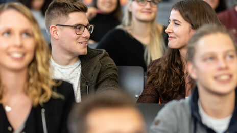 Nahaufnahme mehrerer Studierender in den Sitzreihen eines Hörsaals. Im Fokus eine Studentin und ein Student, die nebeneinander sitzen und sich anlächeln.<br>