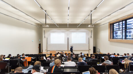 View of a full lecture hall from the back, a lecturer is writing something on the blackboard at the front.