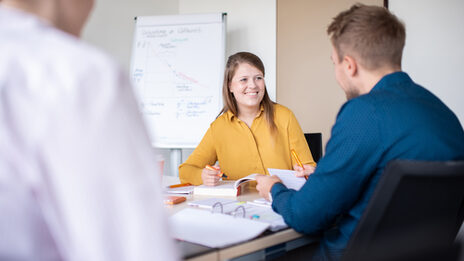 Three students are sitting at a table full of documents. One student looks laughingly in the direction of a fellow student and marks something in the book with a highlighter.