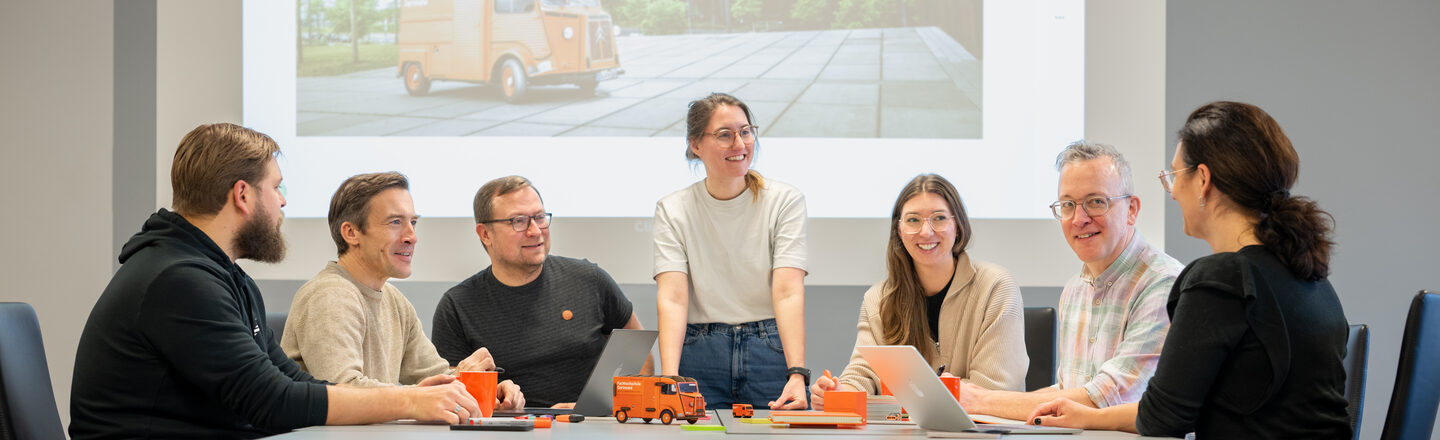 A group of seven people sit around a table while a woman stands looking to the right. A presentation is projected onto the wall in the background. A table with a meeting folder can be seen out of focus in the foreground