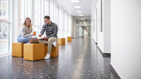 Foto von einer Studentin und einem Studenten. Sie sitzen im Flur in der FH und unterhalten sich. Der Student zeigt auf etwas am Laptop.