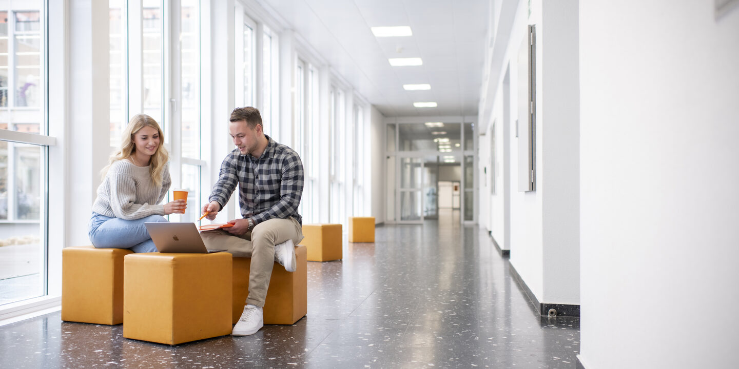 Foto von einer Studentin und einem Studenten. Sie sitzen im Flur in der FH und unterhalten sich. Der Student zeigt auf etwas am Laptop.