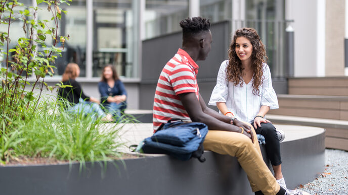 Foto von zwei Studierenden, die im Innenhof sitzen und sich unterhalten. Im Hintergrund zwei weitere sitzende Personen und ein Gebäude. __ Young woman and young man are sitting in the inner courtyard and talking, in the background two other people and a building.