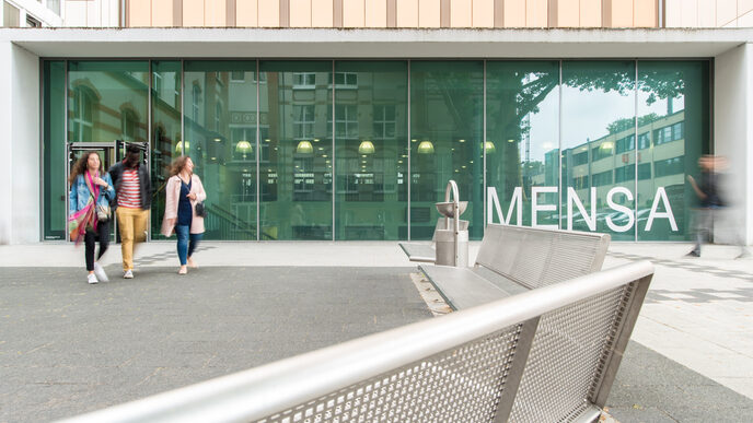 Foto des Mensagebäudes, im Vordergrund Bänke. Vor der Mensa verschwommen vier Personen.__The canteen building, benches in the foreground. Four people blurred in front of the canteen.