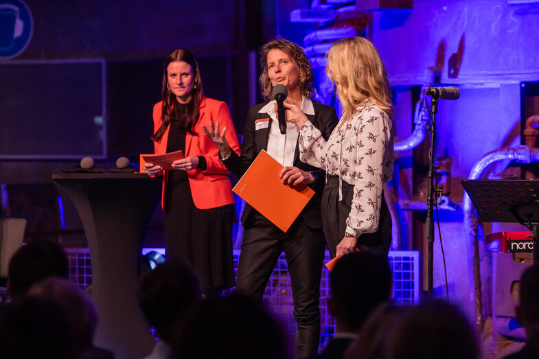 Three people stand on the stage, one holds a certificate and speaks into the microphone.