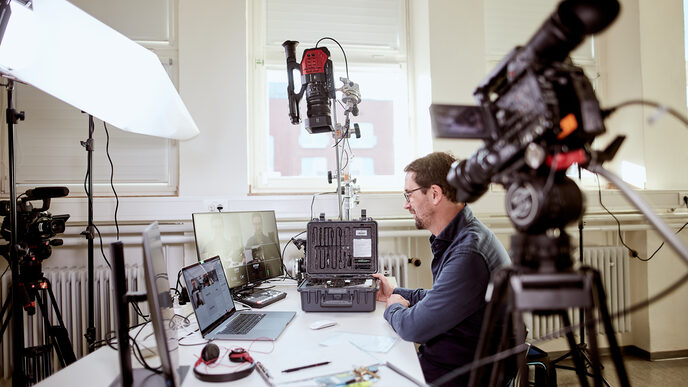 Foto von einem Lehrenden am Schreibtisch, umgeben von diversen Kameraequipment sowie Laptop und Monitoren, um eine online Vorlesung zu halten. __ A prof sits at his desk, surrounded by various camera equipment as well as a laptop and monitors, in order to give an online lecture.