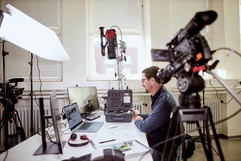 Foto von einem Lehrenden am Schreibtisch, umgeben von diversen Kameraequipment sowie Laptop und Monitoren, um eine online Vorlesung zu halten. __ A prof sits at his desk, surrounded by various camera equipment as well as a laptop and monitors, in order to give an online lecture.