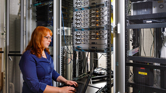 Foto einer Auszubildende vor einem großen Schrank mit Servern, sie bedient einen Laptop. __ <br>A trainee stands in front of a large cabinet with servers and operates a laptop.