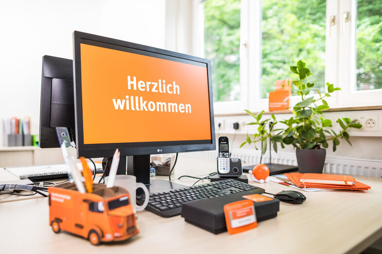 Foto eines Schreibtischs, auf dem sich verschiedene orangefarbene Gegenstände befinden, z.B. ein Block, ein Namensschild und ein Stifthalter in Form des Citroën HY. Auf einem PC-Bildschirm wird <br> „Herzlich Willkommen„ angezeigt. __A desk with various orange objects on it, e.g. a pad, a nameplate and a pen holder in the shape of the Citroën HY. "Welcome" is displayed on a PC screen.