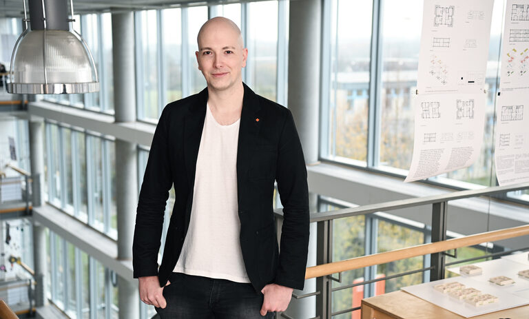 Ein Mensch im schwarzen Sakko steht in einem Gebäude an einem Geländer vor einer Fensterfront.