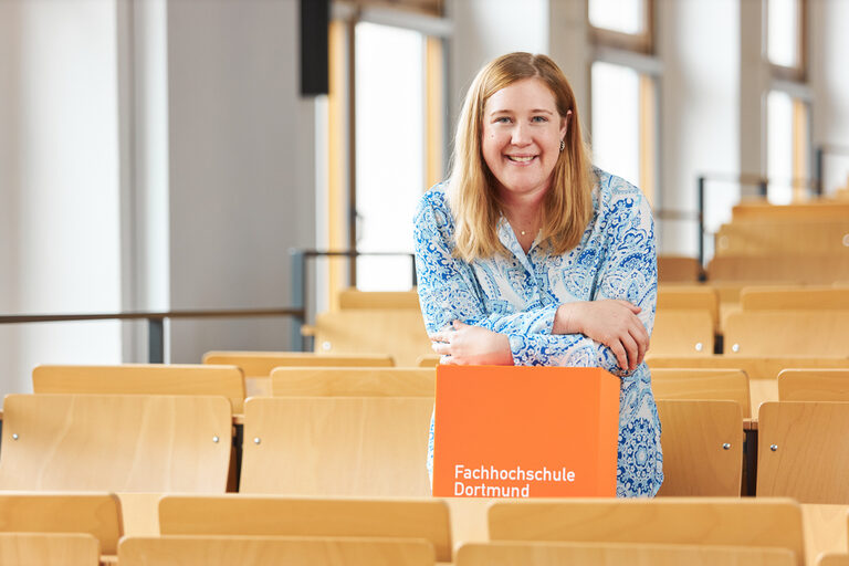 Eine Frau steht in einer Sitzreihe eines Vorlesungsraumes stützt sich auf einem orangenen Würfel ab und lacht.__A woman is standing in a row of seats in a lecture room, leaning on an orange cube and laughing.<br><br>