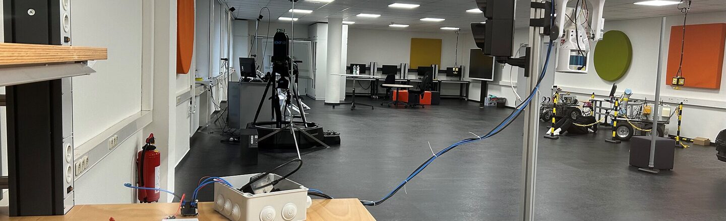A room with a wooden table in the foreground. There are various electronic devices on the table, including a traffic light on a stand. Other technical devices and computer screens can be seen in the background. The room is a laboratory or research room.