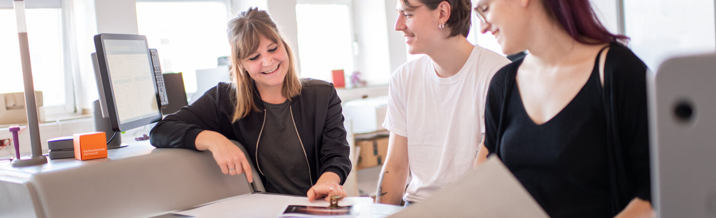 Photo of three people in a printing workshop. The people are discussing and looking at a sketch. They are standing right next to a large printer.