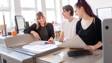 Photo of three people in a printing workshop. The people are discussing and looking at a sketch. They are standing right next to a large printer.