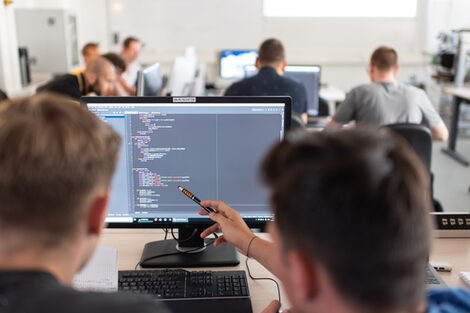 Foto von Studierenden in Rückansicht an Computerarbeitsplätzen. Im Vordergrund zeigt ein Student seinem Sitznachbarn mit einem Stift etwas auf dem Bildschirm.