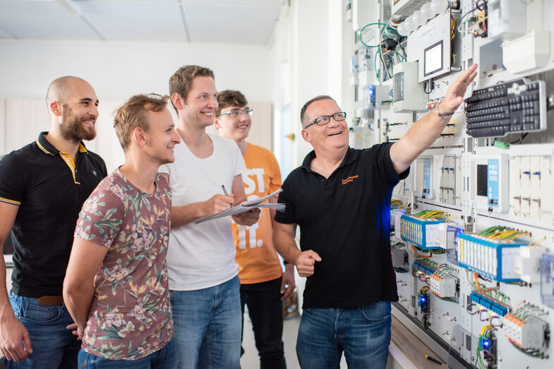 Foto eines Professors, der vor einer Versuchswand der Elektrotechnik steht und auf ein Gerät zeigt. Neben ihm stehen vier lächelnde Studenten, einer von ihnen macht sich Notizen. __ A professor stands in front of an electrical engineering test wall and points to a device. Next to him are four smiling students, one of whom is taking notes.