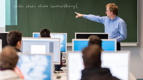 Foto eines Professors, der an der Tafel im Computerraum etwas zeigt. Vor ihm Studierende an Computern.