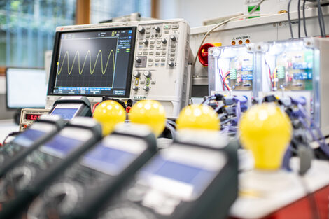 Ein Foto von einem Versuchsaufbau. Im Hintergrund zu sehen sind ein Oszilloskop und zwei von innen beleuchtete Gehäuse mit elektronischer Schaltung. Im Vordergrund unscharf sieht man vier Spannungsmesser und vier LED-Lampen.