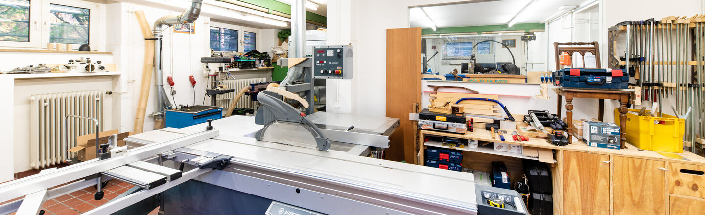 Room shot of part of the wood workshop with large circular table saw and other equipment in the room.