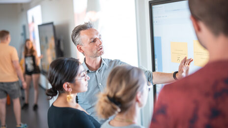 Photo of a group of students standing next to their professor at the whiteboard, who is explaining something to them. In the background, two other students are standing together at a board