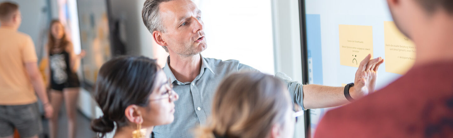 Foto von einer Studierendengruppe, die neben Ihrem Professor am Whiteboard stehen, dieser erklärt Ihnen etwas. Im Hintergrund stehen zwei weitere Studierende zusammen an einem Board