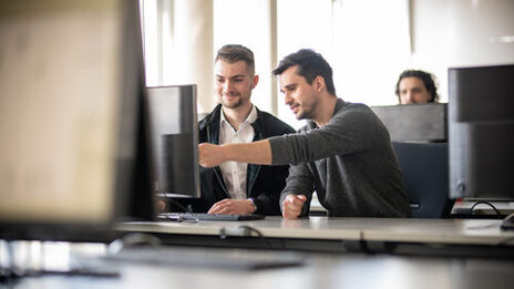 Foto eines Dozenten neben einem Studenten am PC. Der Dozent zeigt dem Studenten etwas auf dem Bildschirm. Im Hintergrund ein weiterer Student an einem Computer.