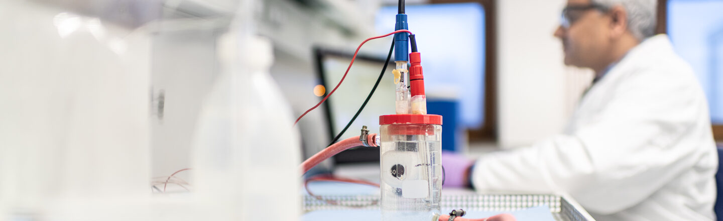 Photo of a test setup in the corrosion laboratory of the Faculty of Mechanical Engineering. Behind it, an employee is sitting at a computer in a blur. The man is wearing a white coat and protective goggles.