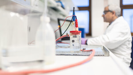 Photo of a test setup in the corrosion laboratory of the Faculty of Mechanical Engineering. Behind it, an employee is sitting at a computer in a blur. The man is wearing a white coat and protective goggles.