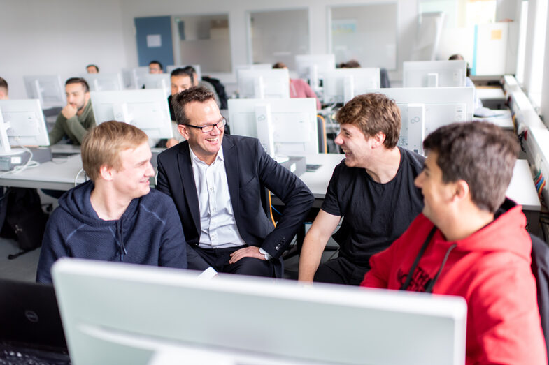 Foto eines Lehrender, der zwischen Studierenden im Computerraum sitzt. Alle schauen den Professor lachend an.