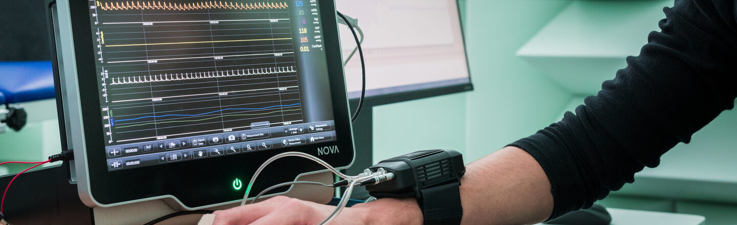 Foto von einer Hand mit angeschlossenem Messgerät auf dem Tisch. Dahinter werden auf einem Monitor Messungen angezeigt beziehungsweise ausgewertet.