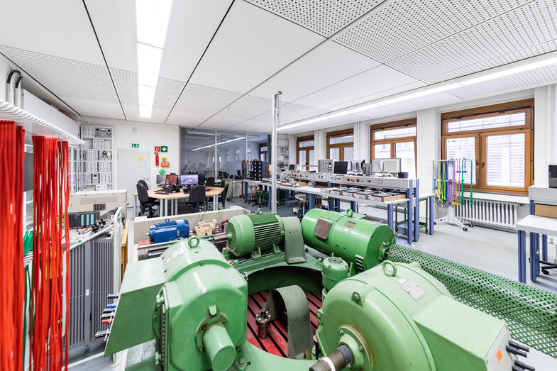 Photograph of the laboratory for power electronics and drive systems with a wide range of laboratory equipment and many devices and machines. In the foreground, electrical machines are mounted on a pedestal. The double tables with experimental setups can be seen in the background. On the right-hand side is the window front facing the inner courtyard of the student workstations.